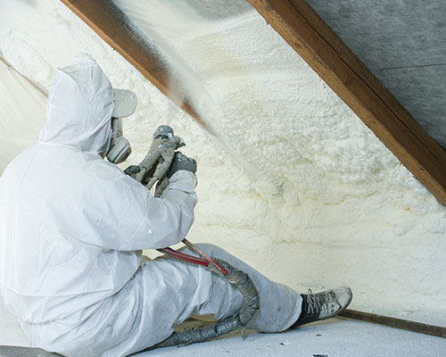 Person spray foaming ceiling in attic wearing protective gear and mask.