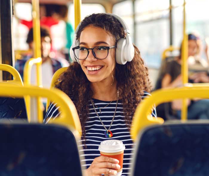 Curly-haired teenager wearing glasses and headphones, sits on an HSR bus and smiles off camera.