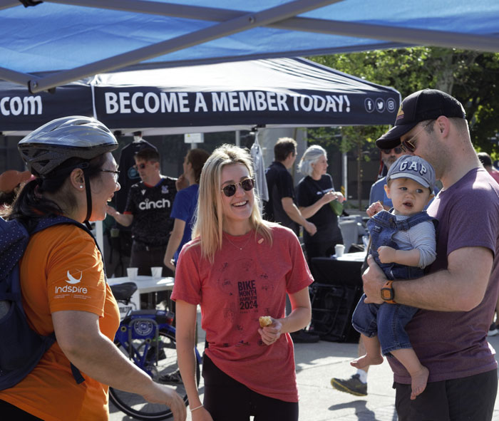 Three adults, one holding a baby, smile and chat at a Bike Month event in Hamilton