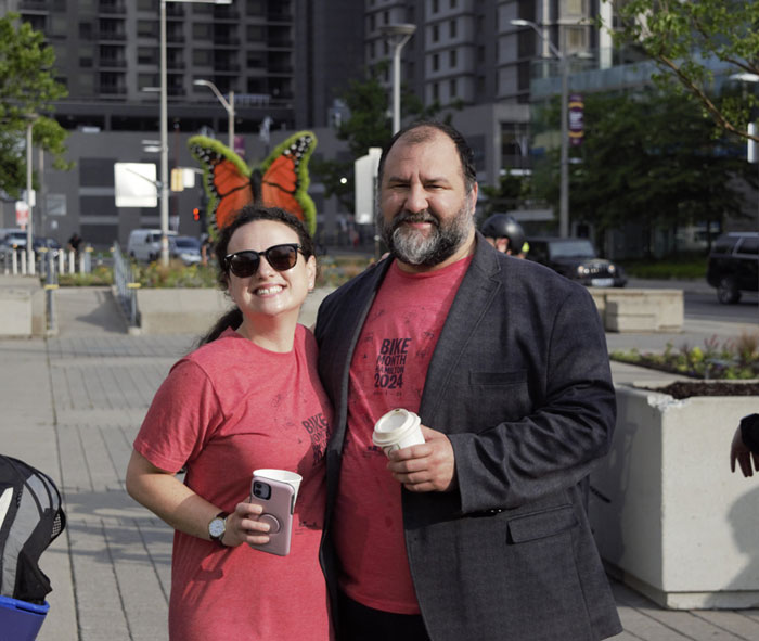 Two people wearing Bike Month Hamilton 2024 t-shirts and holding coffee cups, smile at the camera.