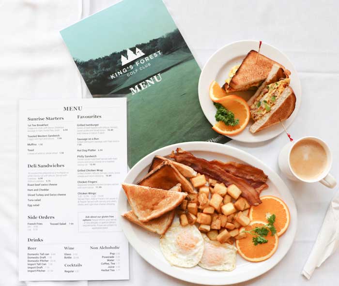 The Kings Forest Gold Club restaurant menu on a table with 2 plates of various breakfast food and a cup of coffee.