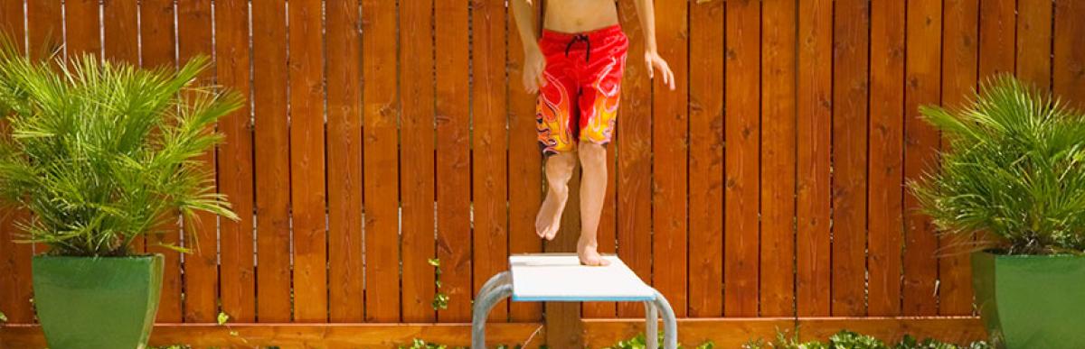 Boy jumping off diving board into pool with wood fence enclosure