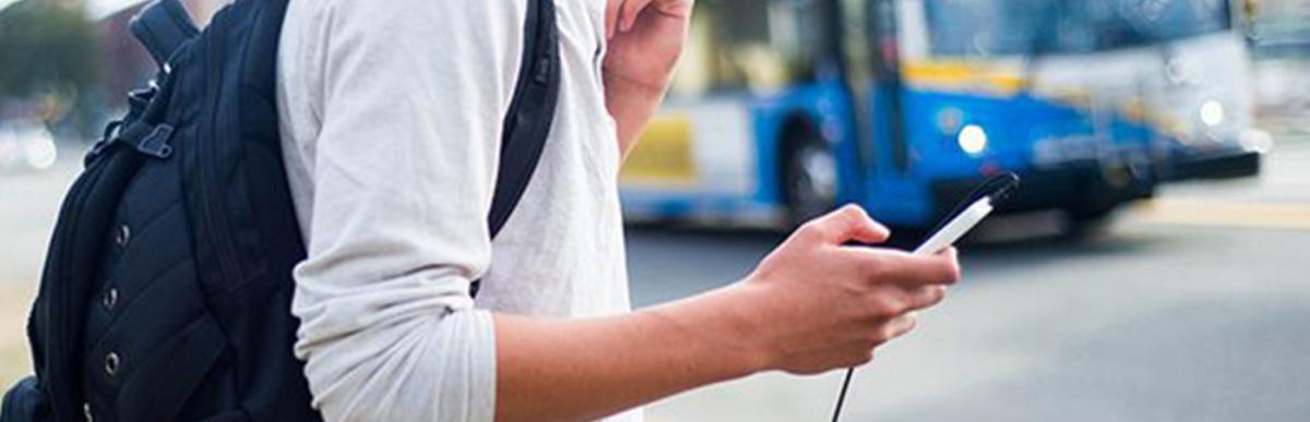 Teenage boy with a backpack, holding a smartphone and waiting for an approaching bus