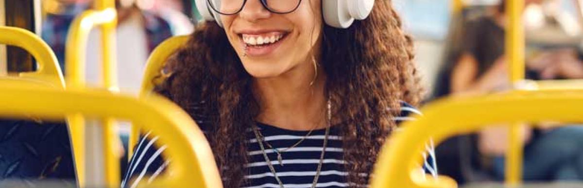 Curly-haired teenager wearing glasses and headphones, sits on an HSR bus and smiles off camera.