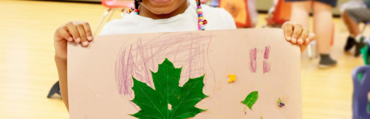 Child smiling at the camera while holding up some artwork they've created with leaves and crayons on paper.