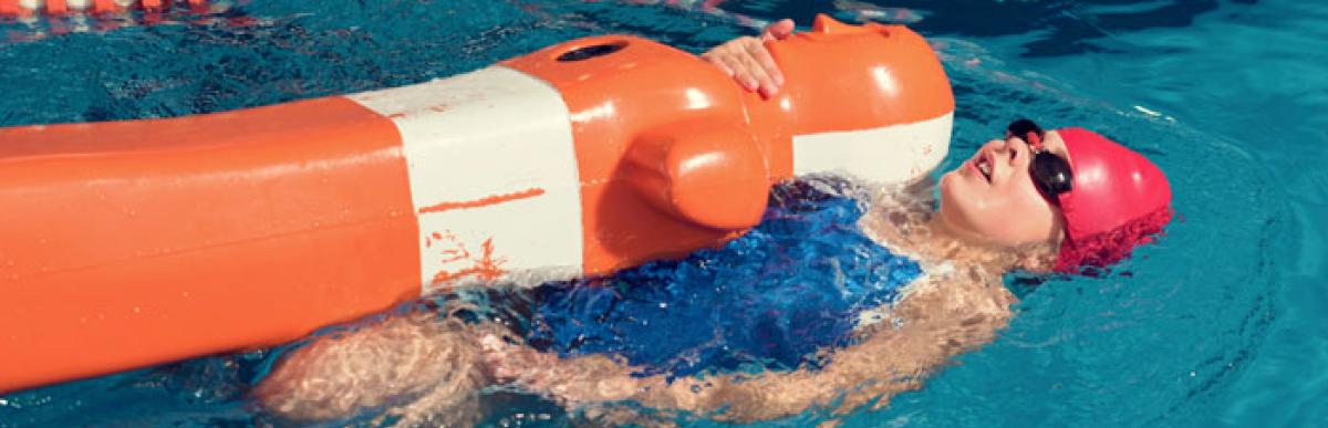 person in a bathing suit, swim cap and goggles, swimming on their back in a pool while holding an orange and white floatation device.