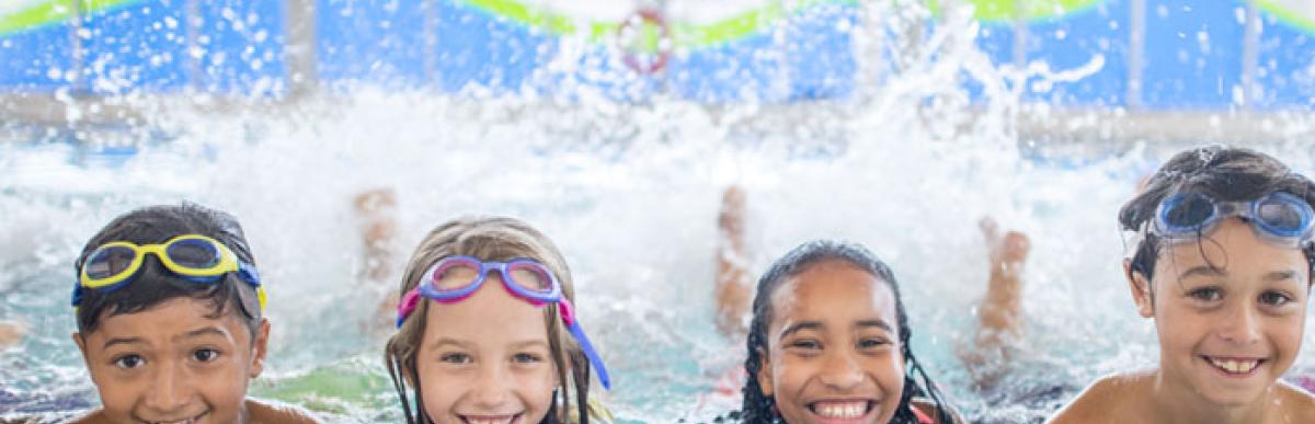 Four children lined up in a row in an indoor pool, smiling at the camera.