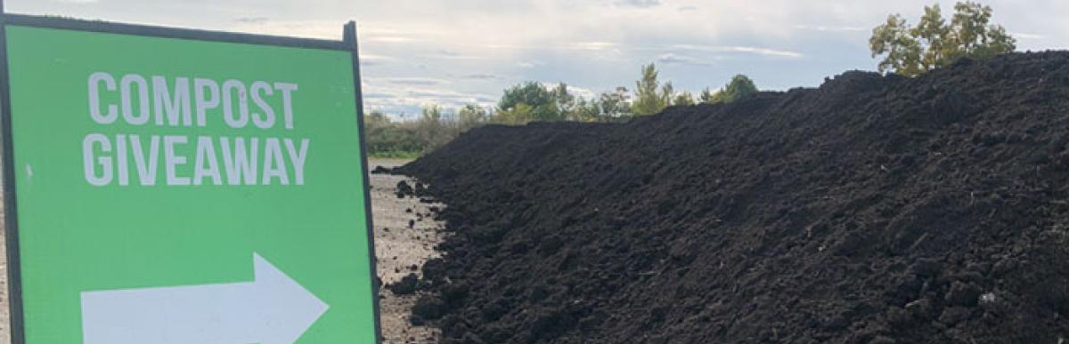 Green sign that says "Compost Giveaway" above an arrow pointing to a large pile of compost beside the sign.