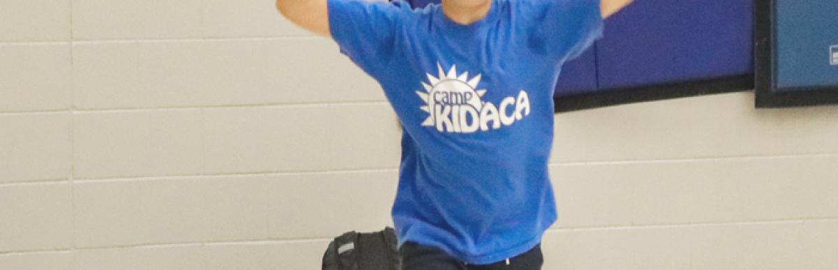 Young boy in a blue Camp Kidaca shirt throws a ball in an indoor gymnasium