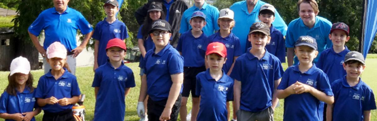 Various kids and golf instructors wearing blue collared golf shirts, standing outside on grass.