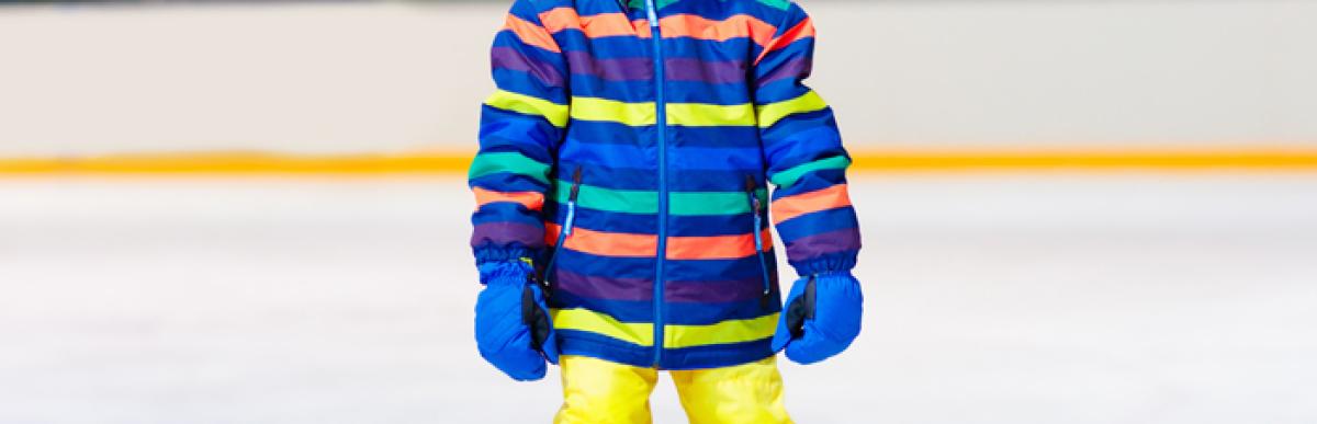 Smiling child in a colourful snowsuit and hocket helmet, wearing skates standing on ice