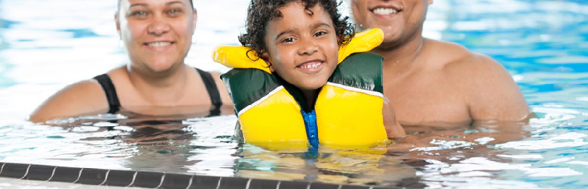 Two smiling adults pose with a smiling child wearing a lifejacket, in an indoor swimming pool