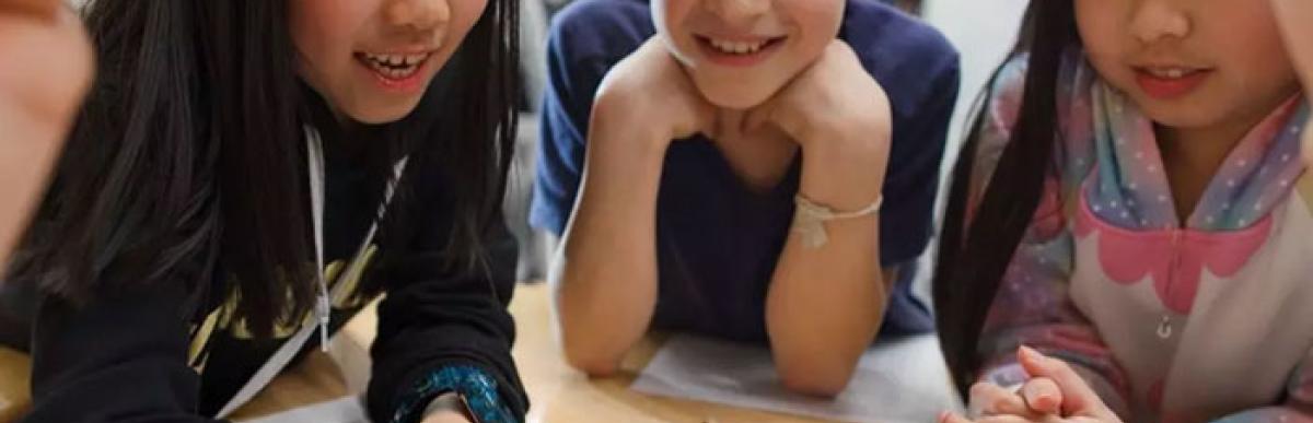 Five smiling children sit facing each other at a desk, working with pencils and paper